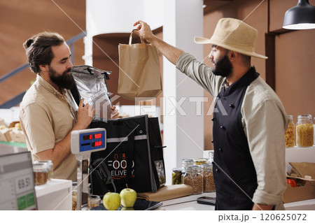 Delivery man is given a paper bag by male vendor. Caucasian courier picks up orders at the neighborhood convenience store and delivers natural, eco-friendly products to customers. 120625072