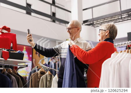 Older people taking pictures in store, comparing blazers on racks with items listed online on app. Senior couple shopping for formal attire during christmas season, looking for festive clothes. Older people taking pictures in store, comparing blazers on racks with items listed online on app. Senior couple shopping for formal attire during christmas season, looking for festive clothes. 120625294