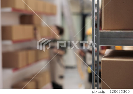 Selective focus of metallic shelves full with carton boxes, in background storehouse workers preparing customers orders. Storage room employees managing parcels transportation, working in warehouse Selective focus of metallic shelves full with carton boxes, in background storehouse workers preparing customers orders. Storage room employees managing parcels transportation, working in warehouse 120625300
