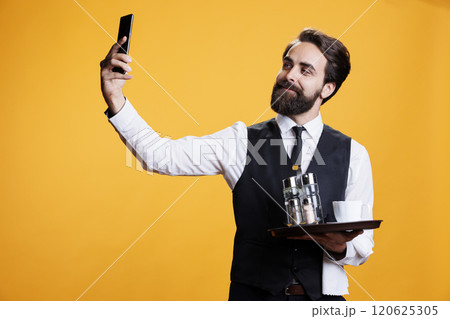 Confident waiter taking selfies in studio, using smartphone camera to capture images of him with restaurant tray in hand. Young man working as butler taking photos against yellow background. 120625305