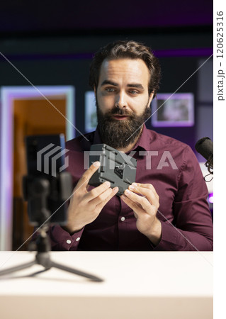 Tech expert filming video review of portable V mount battery using cellphone on tripod, feeling unconvinced of performance. Man analyzing lithium li-ion charging gadget, recording with mobile phone 120625316