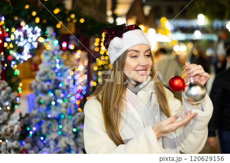 Girl in red Santa hat and with Christmas tree toys in her hands smiles dreamily Girl in red Santa hat and with Christmas tree toys in her hands smiles dreamily 120629156