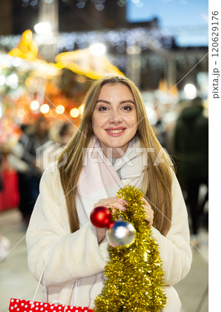 Happy charming girl posing with Christmas balls and tinsel at street fair in evening Happy charming girl posing with Christmas balls and tinsel at street fair in evening 120629176
