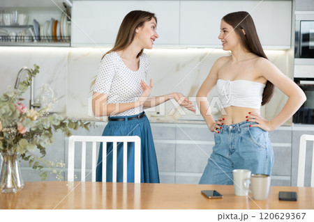 Two young women talking while standing in kitchen 120629367