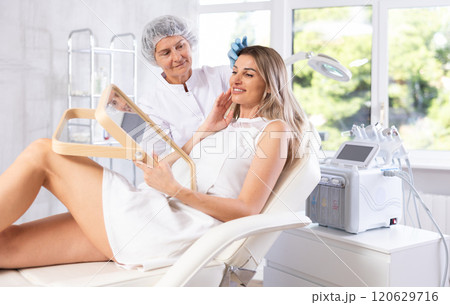 Young woman looking in mirror while mature cosmetologist showing results of facial procedure 120629716