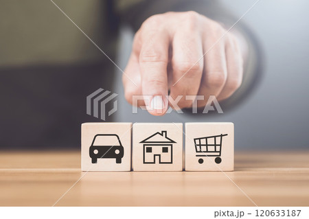 Man considers wooden blocks marked with symbols of a car, house, and airplane, representing choices between investing in a vehicle, home, or travel 120633187