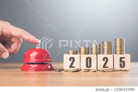Hand presses a red bell next to a stack of coins on a wooden block labeled '2025,' representing financial goals and future planning with a sense of urgency for the upcoming year Hand presses a red bell next to a stack of coins on a wooden block labeled '2025,' representing financial goals and future planning with a sense of urgency for the upcoming year 120633390