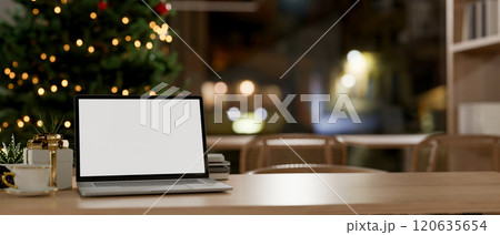 A laptop with a white screen mockup on a table in a room with a Christmas tree in the background. 120635654