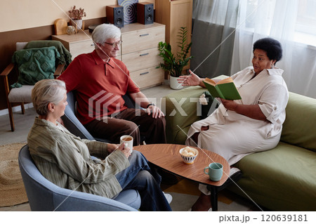 High angle view at group of senior people enjoying book club discussion in cozy living room at retirement community with African American woman reading aloud 120639181