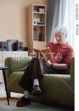 Vertical full length portrait of white haired senior man reading book sitting on sofa at home and enjoying quiet entertainment 120639318