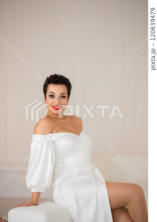 A woman in a white dress is sitting on a white couch. She is wearing a necklace and a red lipstick. A woman in a white dress is sitting on a white couch. She is wearing a necklace and a red lipstick. 120639479