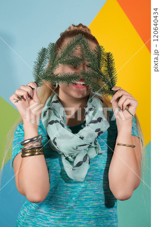 A woman covered her face with Christmas tree branches and smiles. Behind her is a colorful background 120640434