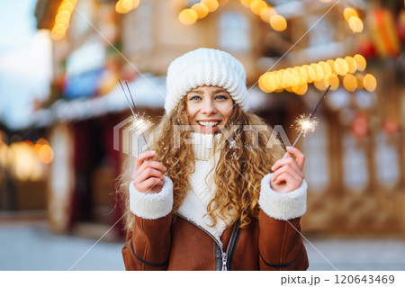 Cheerful young woman holding a sparkler in hand in the winter street. Festive garland lights. Cheerful young woman holding a sparkler in hand in the winter street. Festive garland lights. 120643469