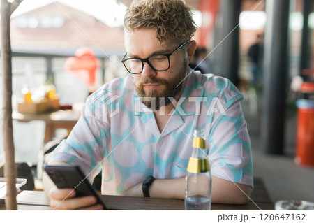 Millennial man with glasses and a patterned shirt sits at an outdoor cafe, focused on his smartphone. Technology and Relaxed summer moments concept Millennial man with glasses and a patterned shirt sits at an outdoor cafe, focused on his smartphone. Technology and Relaxed summer moments concept 120647162