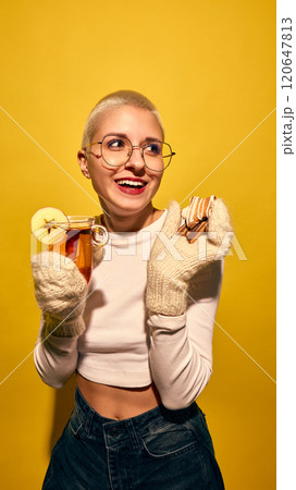 Smiling young woman with short hair, in knitted gloves eating gingerbread cookies and drinking hot cider, against bright yellow background Smiling young woman with short hair, in knitted gloves eating gingerbread cookies and drinking hot cider, against bright yellow background 120647813