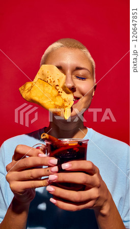 Funny image of young woman with short hair, holding glass of mulled wine with crepe pancake flying to her face, set against red background Funny image of young woman with short hair, holding glass of mulled wine with crepe pancake flying to her face, set against red background 120647851