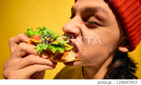Portrait of young man in red beanie hat emotionally eating hot dog with lettuce, mouth covered with mustard, set against bright yellow background Portrait of young man in red beanie hat emotionally eating hot dog with lettuce, mouth covered with mustard, set against bright yellow background 120647852
