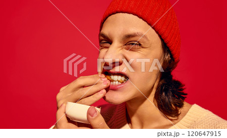 Young emotive man in red beanie hat enjoying sweets, eating donuts with sauce against bright red background 120647915
