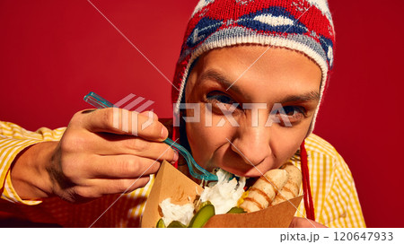 Close-up of emotive young man in winter hat and yellow sweater eating passionately sausages, Bratwurst with pickles and sauerkraut against red background 120647933