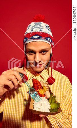 Young guy in stylish winter hat and yellow sweater eating delicious sausages, Bratwurst with pickles and sauerkraut against red background Young guy in stylish winter hat and yellow sweater eating delicious sausages, Bratwurst with pickles and sauerkraut against red background 120647934