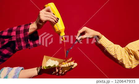 People eating delicious grilled sausages, Bratwurst on carton tray against bright red background. Adding mustard, eating with pickles and sauerkraut People eating delicious grilled sausages, Bratwurst on carton tray against bright red background. Adding mustard, eating with pickles and sauerkraut 120647935