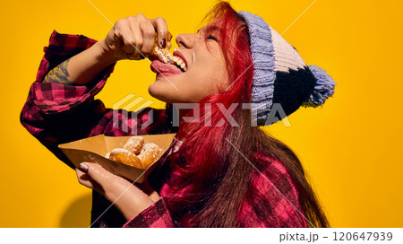 Side view portrait of happy young redhead woman in knitted hat eating mini donuts with her face covered in sugar powder, against yellow background 120647939