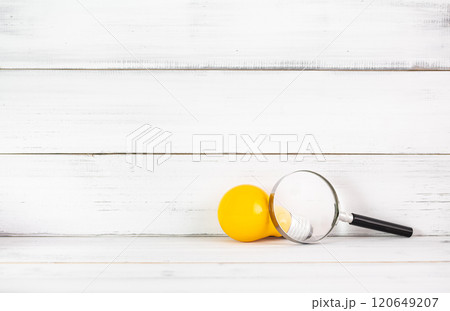 Exploring Bright Ideas: Yellow Light Bulb with Magnifying Glass on Rustic White Background 120649207