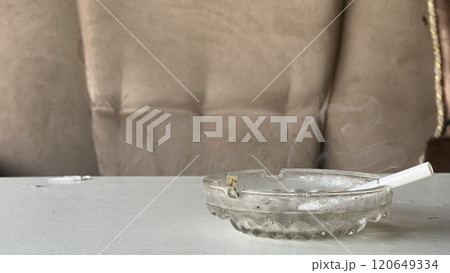 Cigarette in ashtray smoldering on a table near a couch Cigarette in ashtray smoldering on a table near a couch 120649334