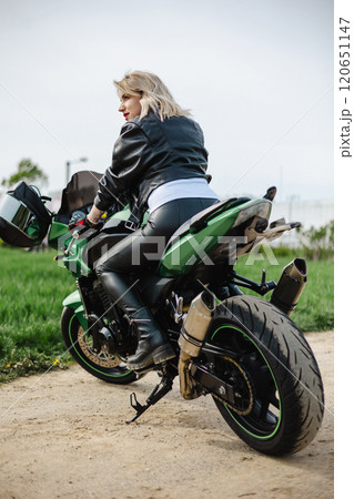 A stylish woman in leather pants and a black jacket sits confidently on a green motorcycle. The A stylish woman in leather pants and a black jacket sits confidently on a green motorcycle. The 120651147