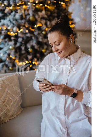 A cheerful woman in a white shirt sits on a cozy couch using her smartphone. Festive lights and a A cheerful woman in a white shirt sits on a cozy couch using her smartphone. Festive lights and a 120651190