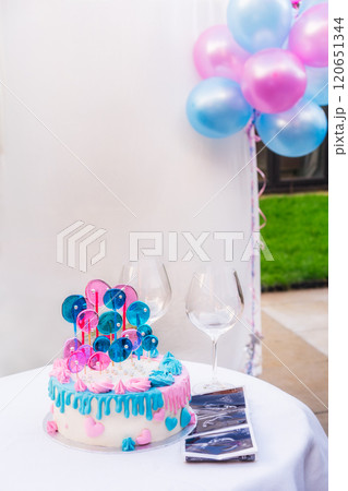 Gender party cake. Boy or girl concept. Baby shower party celebration cake with white chocolate frosting, blue and pink decoration on the white table with glasses and baby ultrasound image print. 120651344