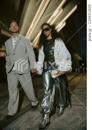 Vertical motion shot of Middle Eastern woman wearing silver trousers holding hands with mature man in classy gray suit, while walking in city street with in night lights in backdrop 120651608