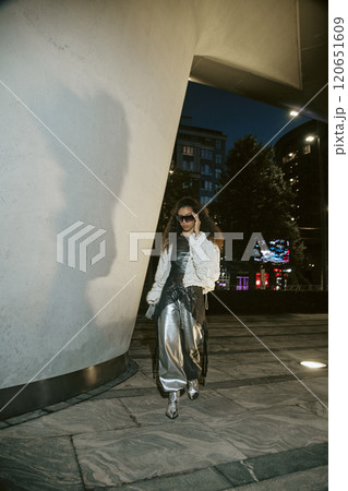 Glamorous Middle Eastern woman adjusting sunglasses walking confidently in silver outfit paired with oversized jacket while posing for paparazzi at night street with city illumination, copy space 120651609