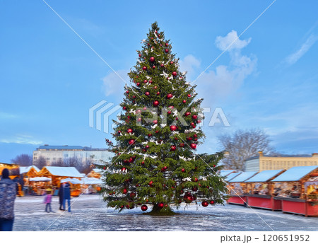 Festively decorated Christmas tree with red ornaments in an outdoor holiday market under clear blue sky 120651952