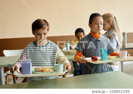 Medium shot of young boy and Central Asian girl on break, holding trays of lunch getting ready to sit down and eat at dining table in school cafeteria Medium shot of young boy and Central Asian girl on break, holding trays of lunch getting ready to sit down and eat at dining table in school cafeteria 120652195