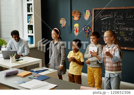 Adorable young students from diverse ethnic backgrounds standing in front of class presenting results of collaborative task to classmates during lesson in modern classroom Adorable young students from diverse ethnic backgrounds standing in front of class presenting results of collaborative task to classmates during lesson in modern classroom 120652306