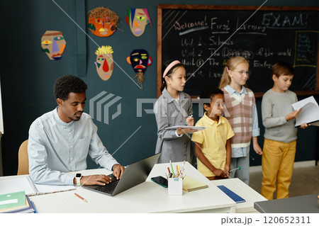 Concentrated African American male teacher typing on laptop sitting at desk, while group of young students standing at blackboard presenting homework results in classroom with blue walls Concentrated African American male teacher typing on laptop sitting at desk, while group of young students standing at blackboard presenting homework results in classroom with blue walls 120652311