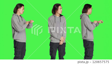 Young male model looks at watch standing against greenscreen backdrop, acting impatient in studio. Asian person feeling frustrated and irritated, waiting for a certain something. Young male model looks at watch standing against greenscreen backdrop, acting impatient in studio. Asian person feeling frustrated and irritated, waiting for a certain something. 120657575