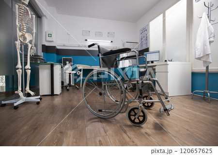 Medical wheelchair placed at center of empty doctors office with a desk and computer for medical consultation. Clinic room is filled with essential healthcare instruments and treatment aids. 120657625