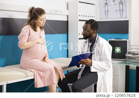 Black doctor and pregnant white woman engage in prenatal consultation in clinic office. Physician talks about medical care plan while expecting mother listens with her hands resting on pregnancy belly 120657628