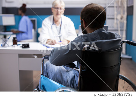 Selective focus of young man in wheelchair talking with older female physician in clinic office. Rearview closeup of bearded male patient in medical consultation with elderly doctor. 120657755