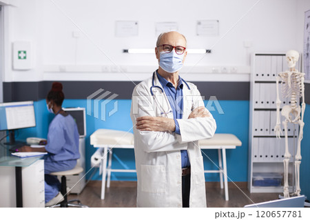 Caucasian retired doctor wearing face mask is standing confidently in modern clinic with arms crossed. Masked senior male physician in medical office looking at camera, ready for patients. Caucasian retired doctor wearing face mask is standing confidently in modern clinic with arms crossed. Masked senior male physician in medical office looking at camera, ready for patients. 120657781