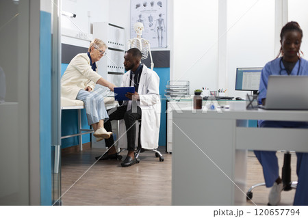 Black doctor engages in conversation with retired white woman using clipboard to outline treatment plan. Caucasian old patient asking questions as physician listens attentively in hospital room. Black doctor engages in conversation with retired white woman using clipboard to outline treatment plan. Caucasian old patient asking questions as physician listens attentively in hospital room. 120657794