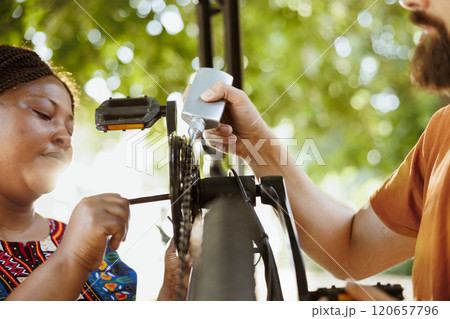 Young multiracial pair servicing and maintaining bike chain and pedals by using specialised equipment. Sports-loving boyfriend and girlfriend greasing and securing bicycle parts in home yard. Young multiracial pair servicing and maintaining bike chain and pedals by using specialised equipment. Sports-loving boyfriend and girlfriend greasing and securing bicycle parts in home yard. 120657796