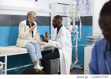 Caucasian pensioner patient sits on hospital bed as african american doctor advises on usage of medicine for her recovery. Friendly physician giving prescribed botte of pills to old white woman. Caucasian pensioner patient sits on hospital bed as african american doctor advises on usage of medicine for her recovery. Friendly physician giving prescribed botte of pills to old white woman. 120657797