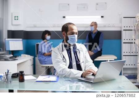 Male general practitioner sits in hospital office, using laptop and planning treatments for his patients during appointments. Doctor checking emails and preparing medical consultations in clinic. 120657802
