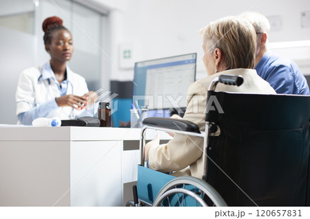 Selective focus of elderly woman in wheelchair consulting with black doctor about post treatment care and health goals. African american female physician consulting with caucasian senior couple. Selective focus of elderly woman in wheelchair consulting with black doctor about post treatment care and health goals. African american female physician consulting with caucasian senior couple. 120657831