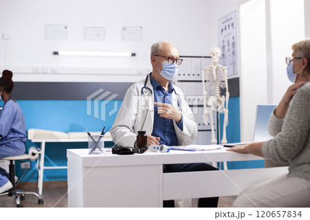 Old male doctor sits across senior white woman in clinic office, discussing checkup results, both wearing face masks for safety. Elderly medic consulting with female pensioner patient in hospital. Old male doctor sits across senior white woman in clinic office, discussing checkup results, both wearing face masks for safety. Elderly medic consulting with female pensioner patient in hospital. 120657834