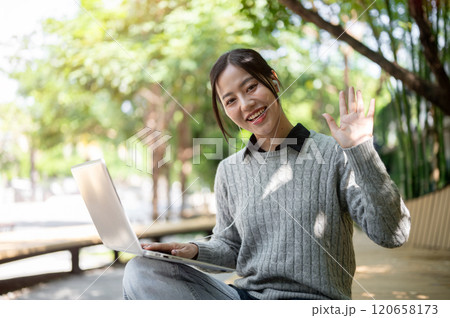 A beautiful Asian woman sits on a park bench with her laptop, smiles warmly and waves at the camera. A beautiful Asian woman sits on a park bench with her laptop, smiles warmly and waves at the camera. 120658173