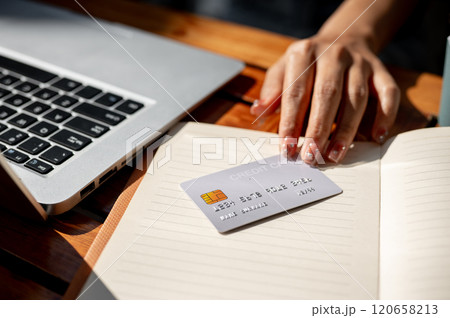 A close-up of a woman's hand holding a credit card on a table, paying bills online through laptop. A close-up of a woman's hand holding a credit card on a table, paying bills online through laptop. 120658213